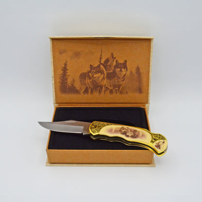 geöffnete helle Geschenkbox mit Wölfen im Deckel, darauf Klappmesser, heller Griff mit Wölfen und Verzierungen, aufgeklappt