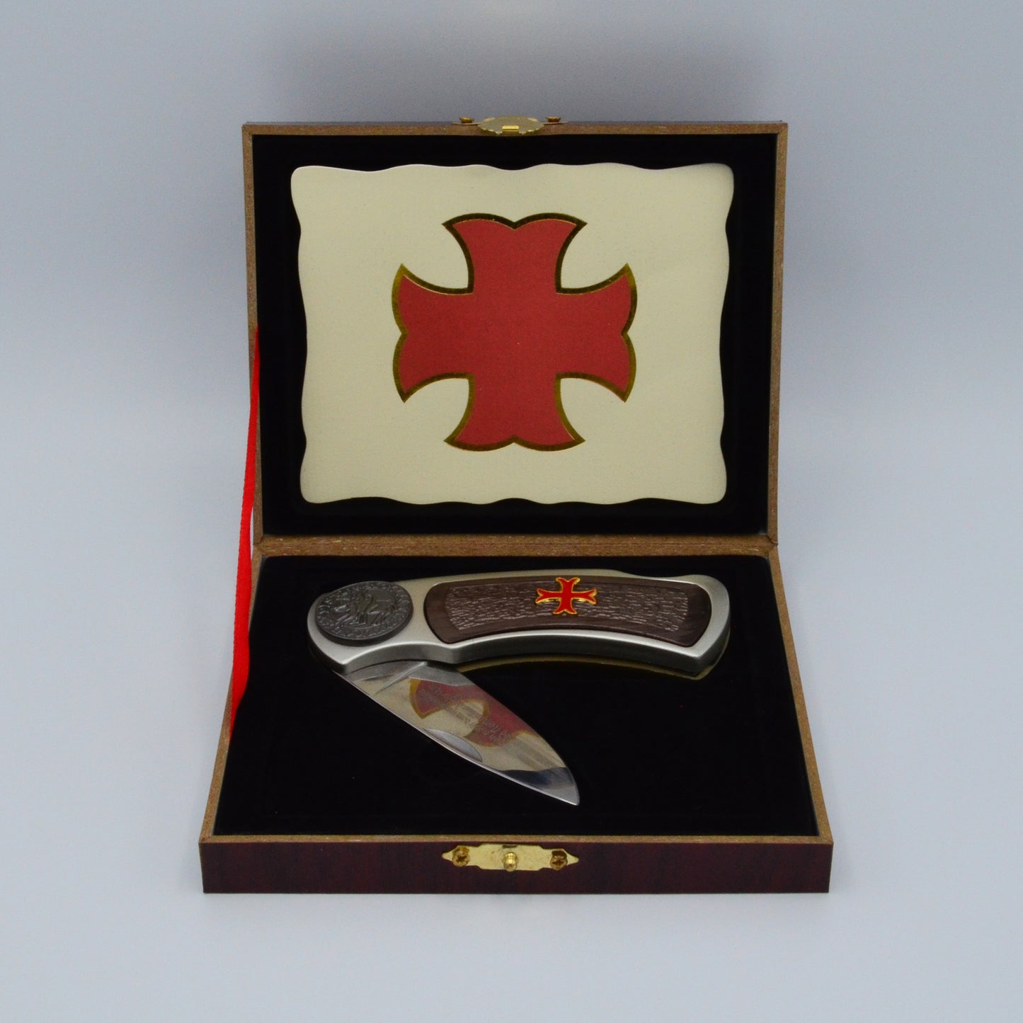 dunkle Geschenkbox, Templerkreuz im Deckel, hält rotes Band, darin Klappmesser mit dunklen Applikationen und Templerkreuz im Griff, Klinge mit Gravur sichtbar