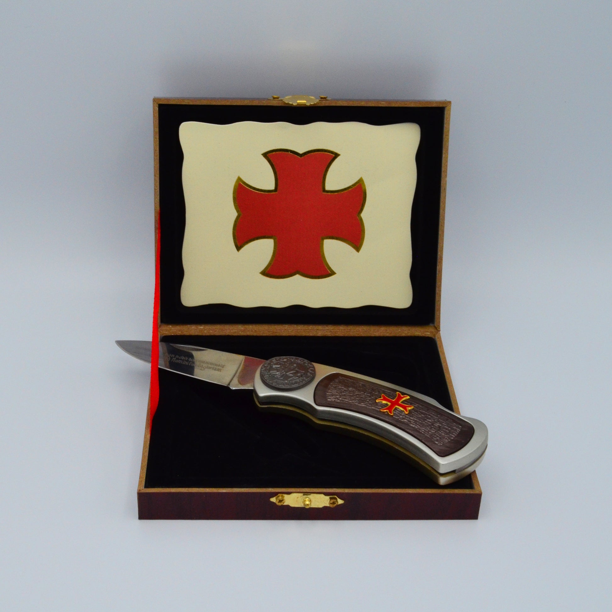 dunkle Geschenkbox, Templerkreuz im Deckel, hält rotes Band, darauf liegt Klappmesser mit dunklen Applikationen und Templerkreuz im Griff aufgeklappt,  Klinge mit Gravur