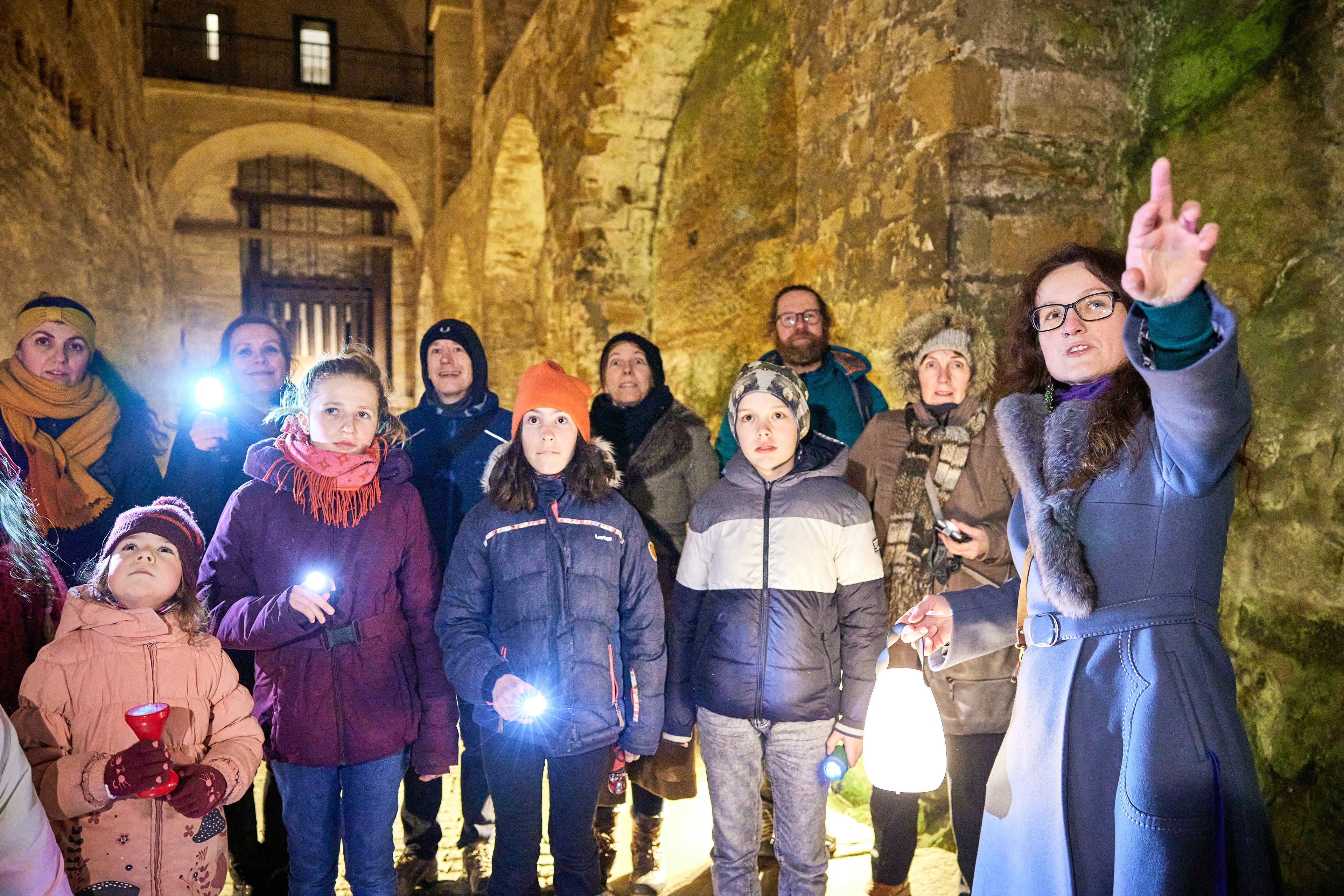 Gruppe Kinder steht mit Gästeführerin und Taschenlampen in dunklem historischen Gemäuer. Gästeführerin zeigt und erklärt etwas.